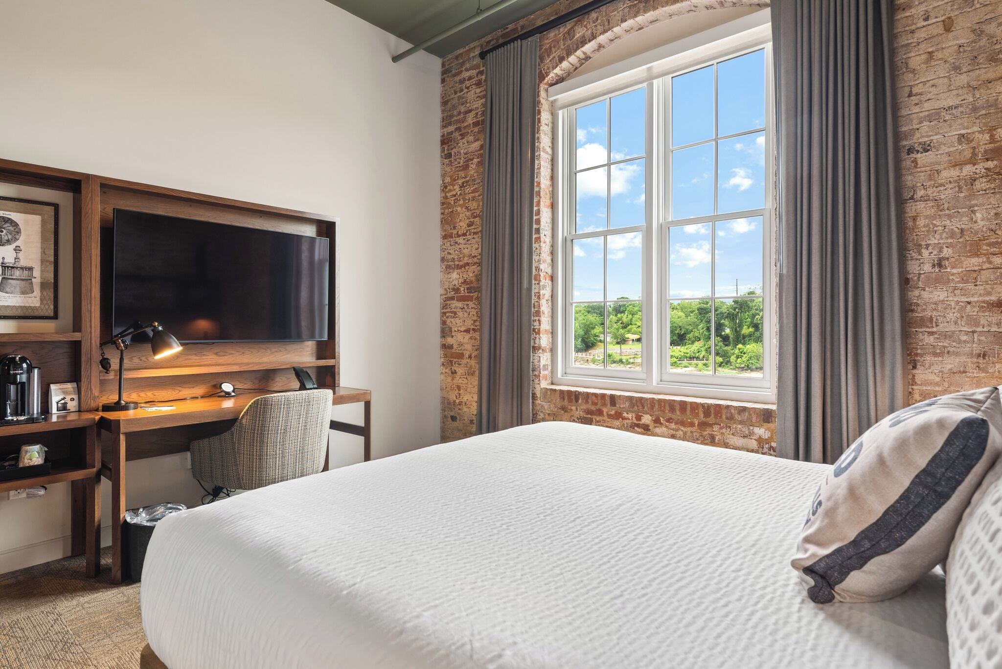 Quarto de hotel com uma cama grande, uma escrivaninha com computador, uma cadeira, televisão e janela com vista para o verde.