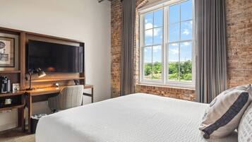 A hotel room with a large bed, a desk with a computer, a chair, a TV, and a window with a view of greenery.