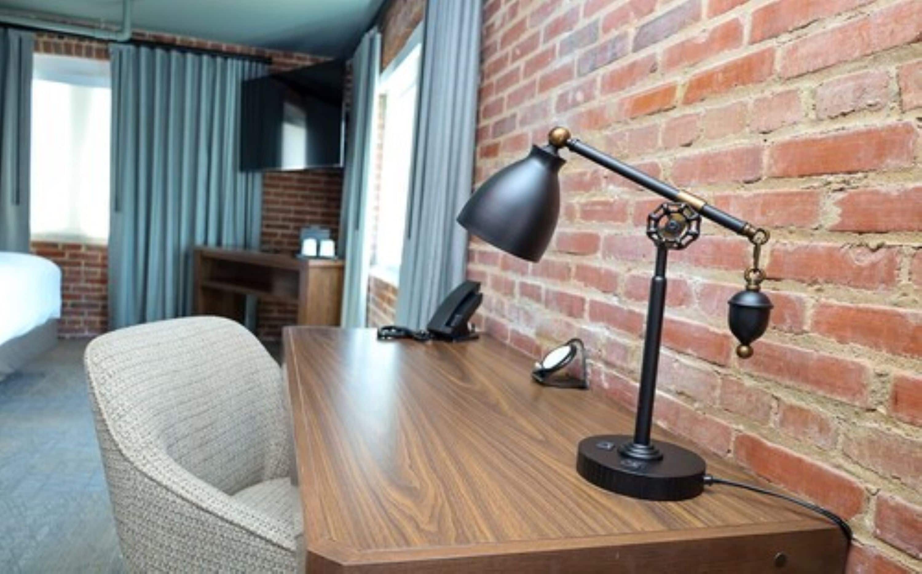 A hotel room with a wooden desk, a chair, a desk lamp, a telephone, and a brick wall.