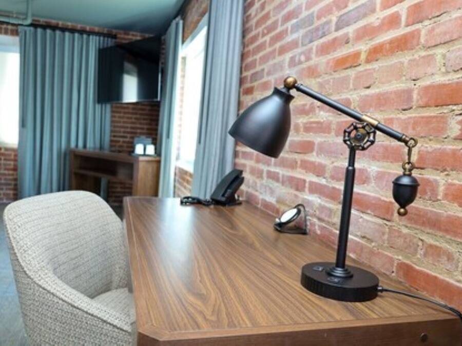 A hotel room with a wooden desk, a chair, a desk lamp, a telephone, and a brick wall.