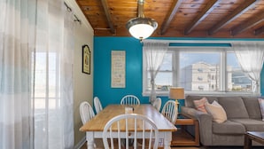 A dining area with a wooden table and white chairs, a blue accent wall, and a view of buildings through a window. - Book Shifting Sands | Sandbridge Beach Vacation Rental (Virginia Beach)