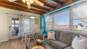 A cozy living room with a wooden ceiling, a gray sofa, a dining table with chairs, and a view through large windows. - Book Shifting Sands | Sandbridge Beach Vacation Rental (Virginia Beach)