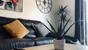 Un salon moderne avec un canapé en cuir noir, une table basse en bois, une plante en pot, une horloge murale et un tableau abstrait aux couleurs vives.