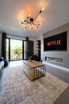 Ein modernes Wohnzimmer mit großem Flachbildfernseher, Couchtisch aus Holz, Kamin und einem Balkon mit Vorhängen.