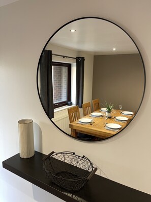 A dining area with a round mirror reflecting the room, a wooden table set with plates and glasses, a potted plant, and a black basket on a black shelf.