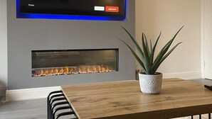 Un salon moderne avec un téléviseur à écran plat fixé au mur, affichant le logo « NETFLIX », une cheminée en dessous, une table basse en bois et une plante en pot.