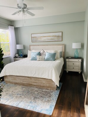 A bedroom with a bed, nightstands, a ceiling fan, a window with curtains, and a painting on the wall.