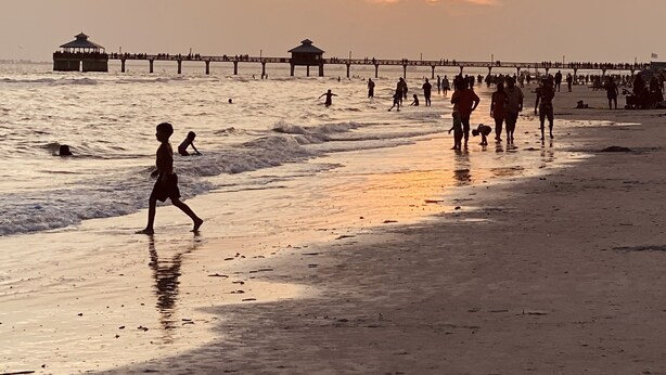 A beach scene with a pier extending into the water, people walking along the shore, and a sunset in the background. - Location, Location, Location (Fort Myers)