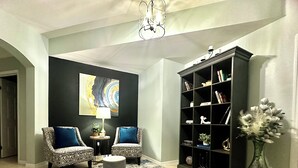 A living room with two patterned armchairs, a small round ottoman, a blue and white rug, a bookshelf, and a vase with flowers. - Location, Location, Location (Fort Myers)