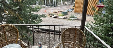 A balcony with wicker chairs and a small table, overlooking a forested area with a dirt path and a wooden structure.