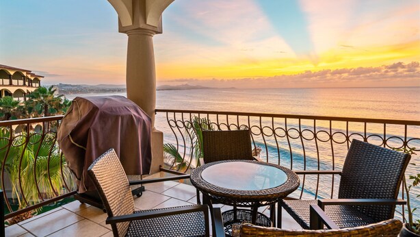 A balcony with a view of the sea at sunset, featuring a covered grill, wicker chairs, and a glass table.