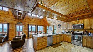 A cabin interior with wooden walls, a kitchen with stainless steel appliances, a fireplace, and a large window overlooking a forested area. - New Construction Mountain Views, Hot Tub and Theater (Pigeon Forge)