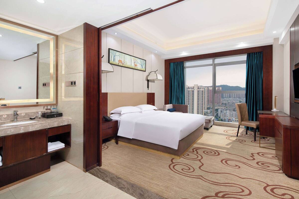 A hotel room with a bed, a desk, a chair, a TV, a large window with curtains, a bathroom with a mirror and sink, and a city view.