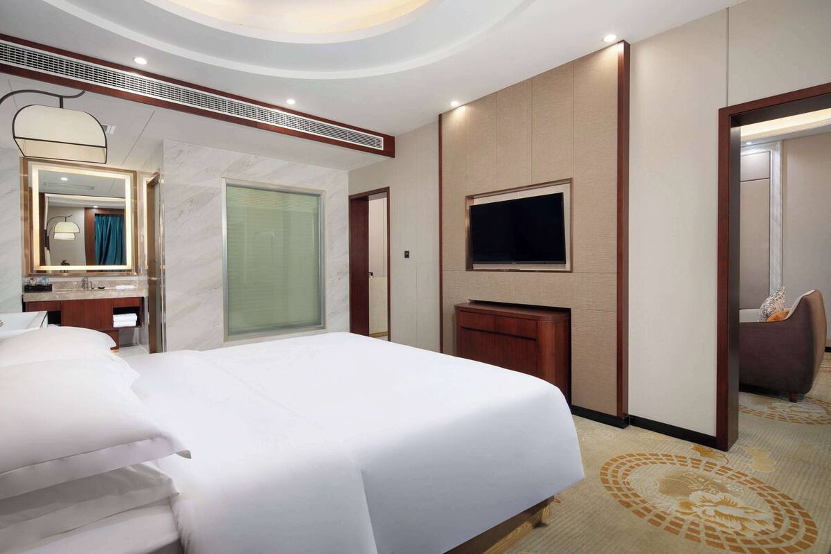A hotel room with a large bed, a TV mounted on the wall, a bathroom with a mirror and vanity, and a carpeted floor.