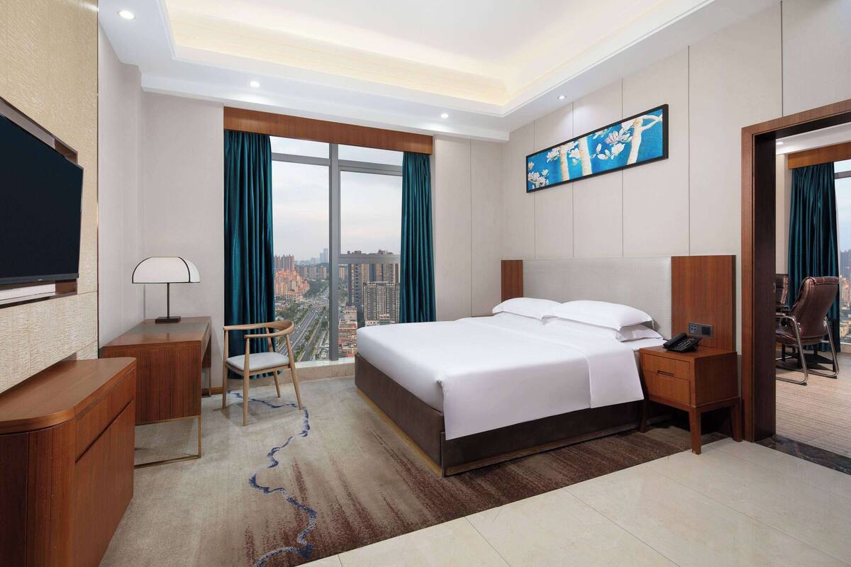 A hotel room with a large bed, a desk, a chair, a TV, and a view of the city.