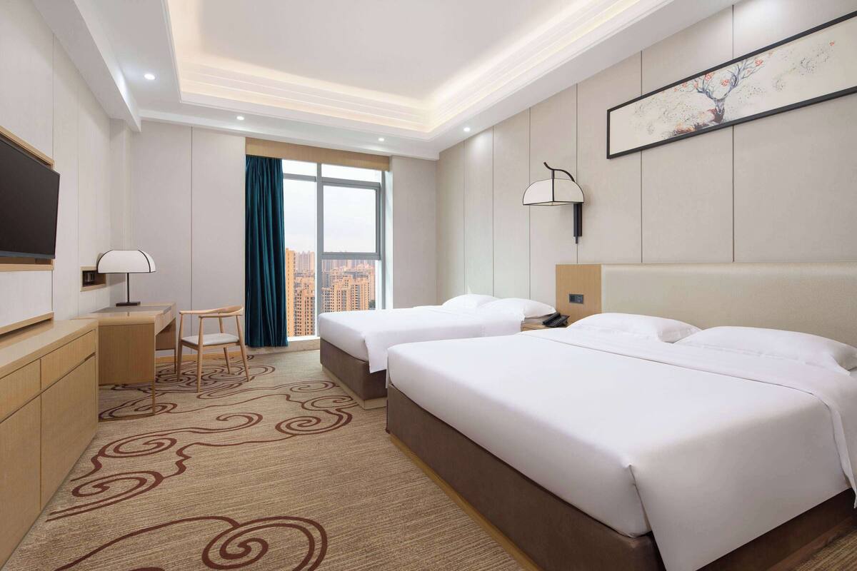 A hotel room with two beds, a desk, a chair, a TV, and a large window with a view of the city.