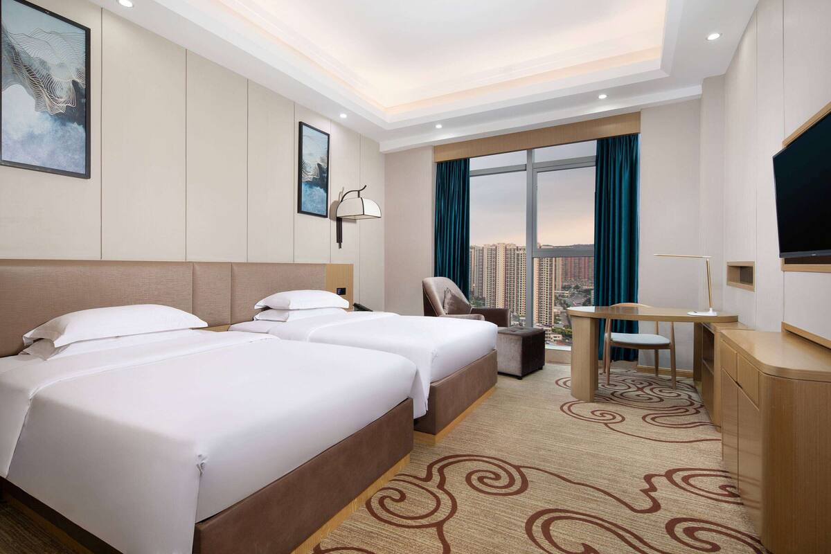 A hotel room with two beds, a desk, a chair, a TV, and a large window with a city view.