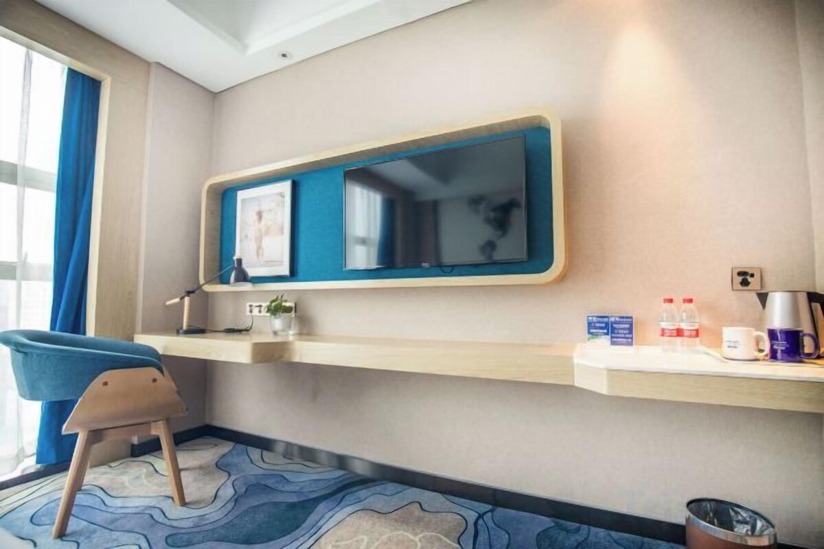 A hotel room with a desk, chair, and a large mirror. There is a small shelf with toiletries and a trash bin.