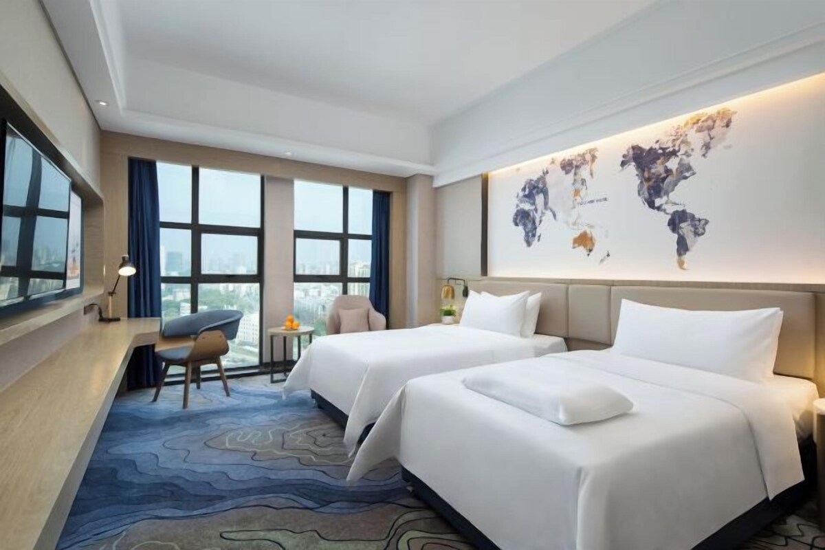 A hotel room with two beds, a desk, a chair, a world map on the wall, and large windows.