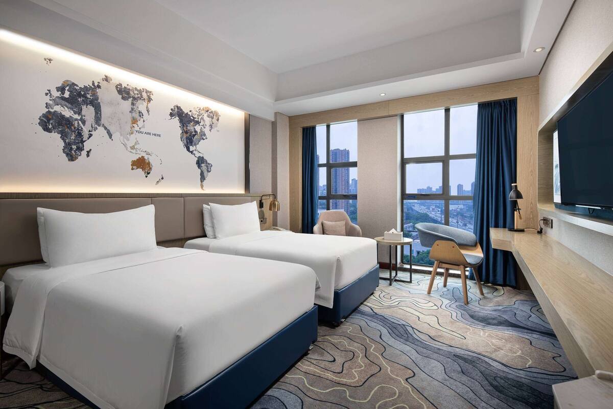 Hotelkamer met twee bedden, een bureau, een stoel, een wereldkaart aan de muur en een groot raam met uitzicht over de stad.