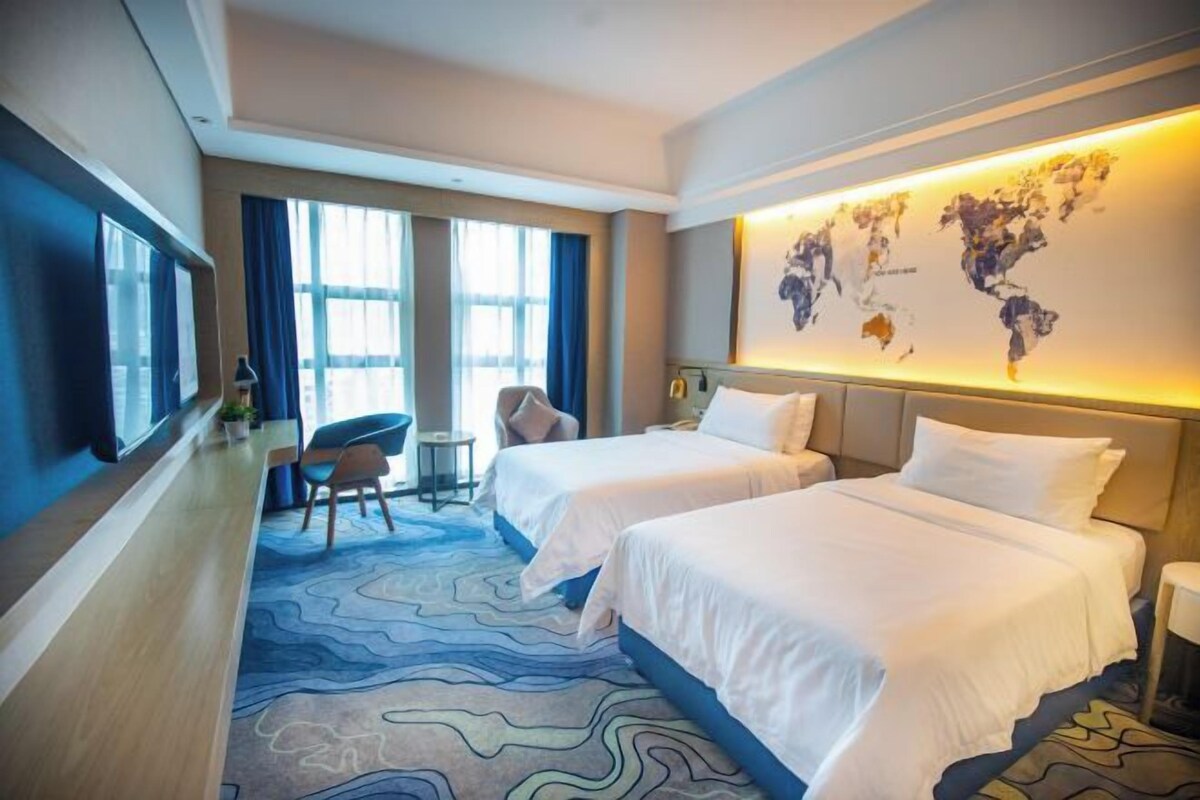 Quarto de hotel com duas camas, uma escrivaninha, uma cadeira, um mapa-múndi na parede e uma janela grande.