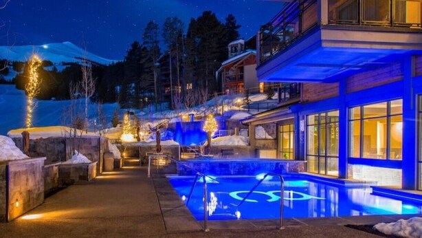 En moderne bygning med poolområde, store vinduer og et snedækket bjerg i baggrunden.