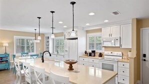 Una cocina amplia con muebles blancos, una isla central, iluminación colgante y vistas al exterior a través de grandes ventanales.
