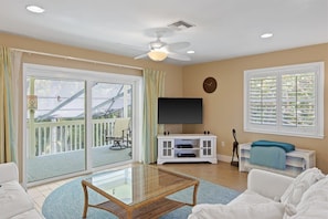 A living room with a glass coffee table, white sofas, a flat-screen TV, a ceiling fan, and a balcony with a view.
