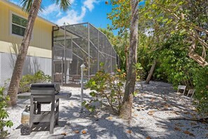 A covered outdoor area with a grill, a small table, and chairs, surrounded by trees and a building.