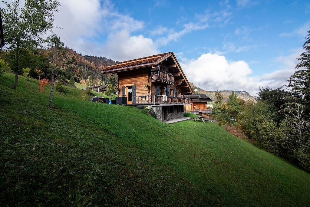 A wooden chalet with a balcony, set on a hillside with a view of a forested mountain range.