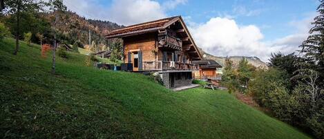 A wooden chalet with a balcony, set on a hillside with a view of a forested mountain range.