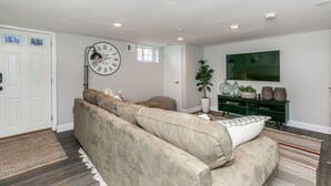 Una sala de estar con un sofá color beige, un reloj de pared grande, un mueble de entretenimiento verde y una planta en maceta.