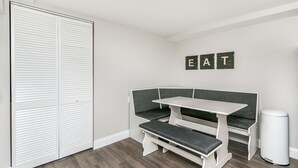 Habitación con un banco blanco, una mesa y un cubo de basura. Hay un armario con puertas correderas y un letrero en la pared que dice “EAT”.