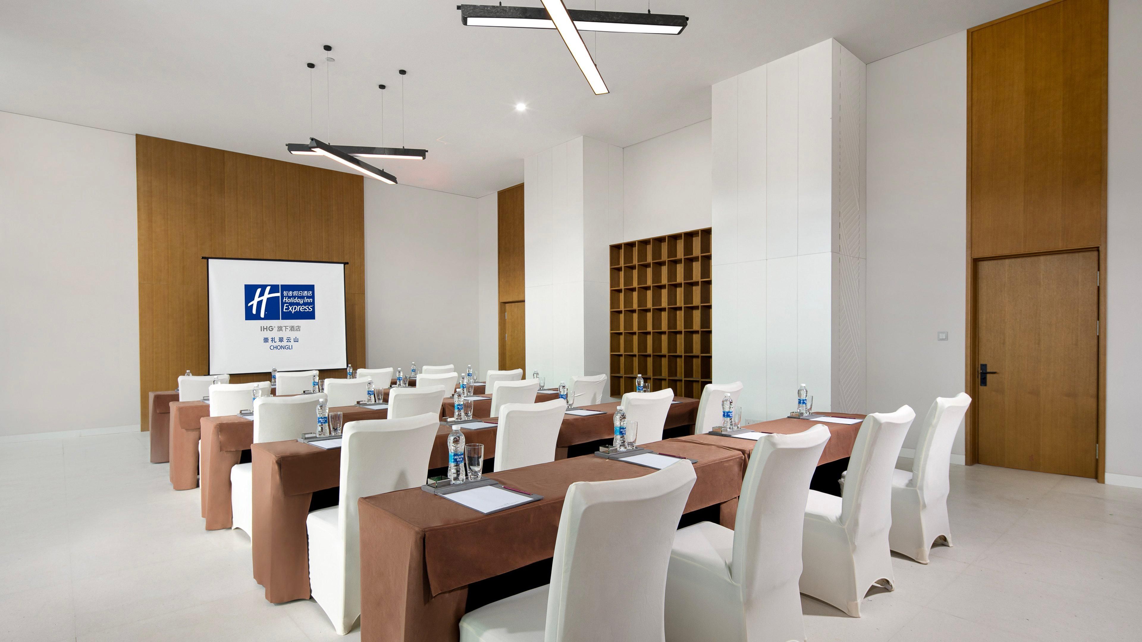 A conference room with multiple tables, white chairs, and a projection screen displaying a logo.