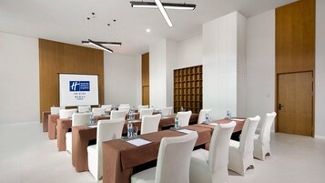 Un salón de conferencias con varias mesas, sillas blancas y una pantalla de proyección que muestra un logotipo.