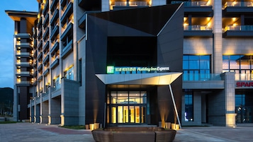 A modern hotel entrance with a Holiday Inn Express sign, a large outdoor seating area, and a multi-story building with balconies.