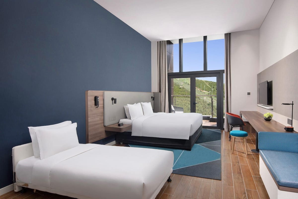 Habitación de hotel con dos camas, un escritorio, un sofá, un televisor y un ventanal con vistas a un paisaje verde.