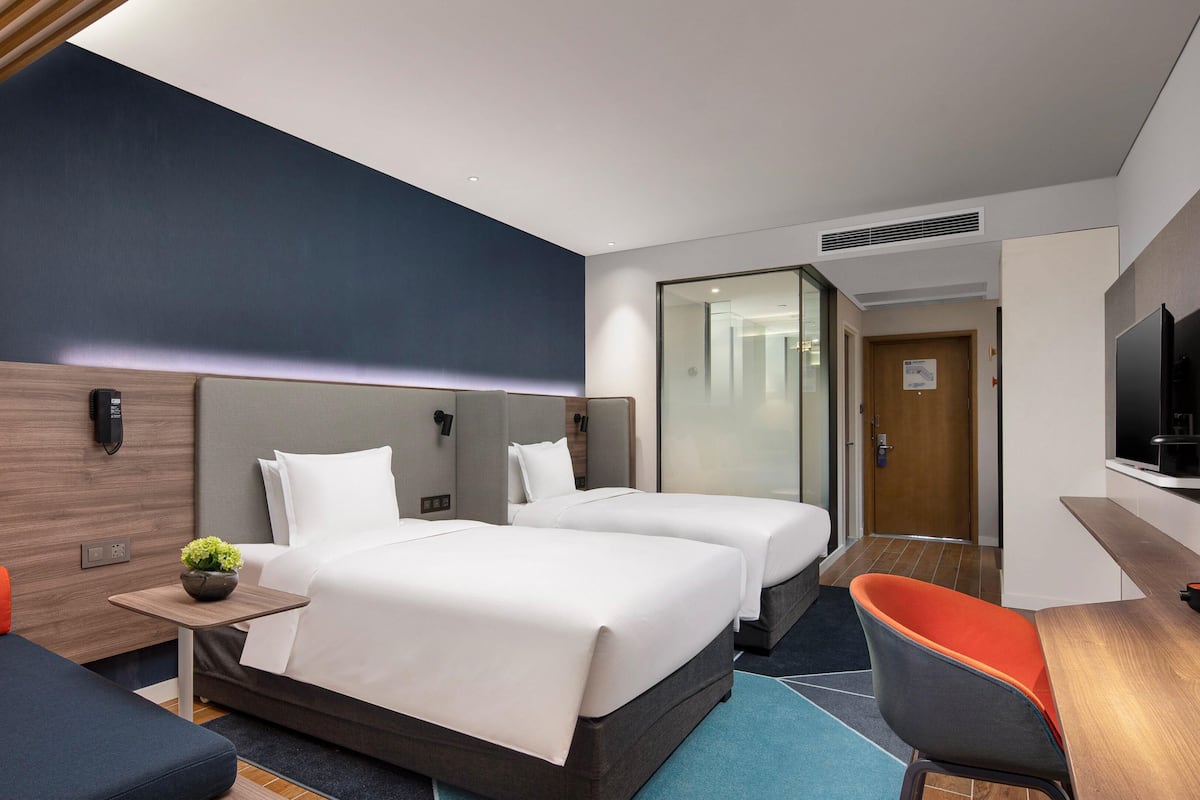 Habitación de hotel con dos camas, un escritorio, un televisor y una zona de estar con una planta.