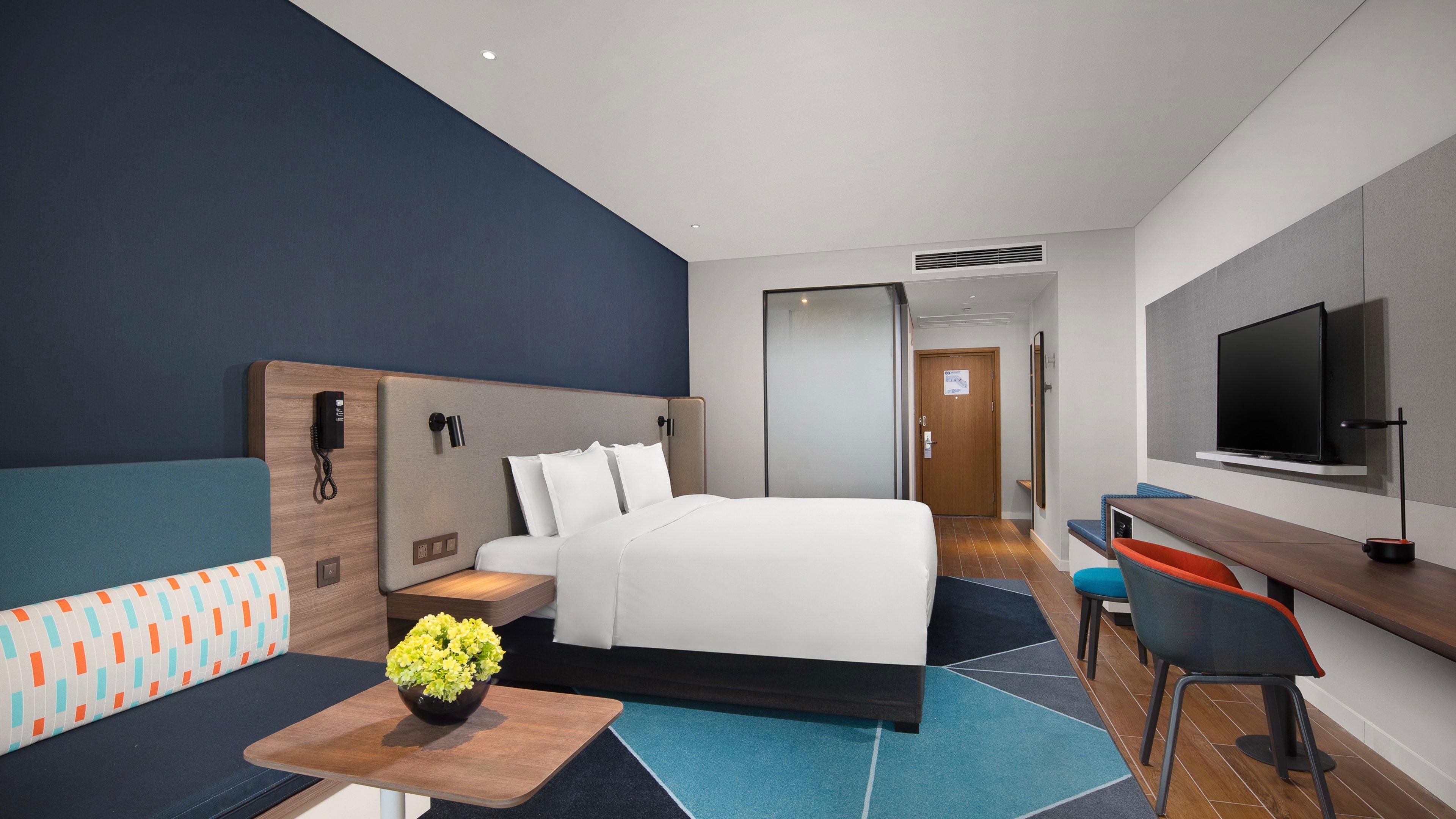 A hotel room with a bed, a desk, a TV, a sofa, and a small table with flowers.