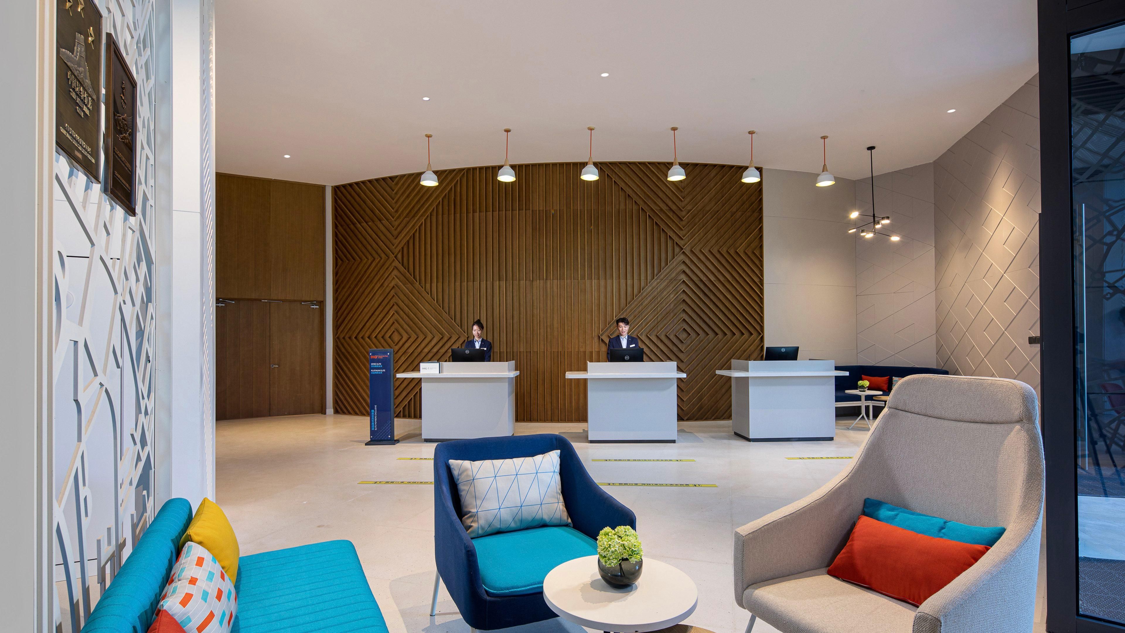 A modern reception area with two reception desks, a blue and a grey chair with cushions, a small table with a vase, and a wooden patterned wall.