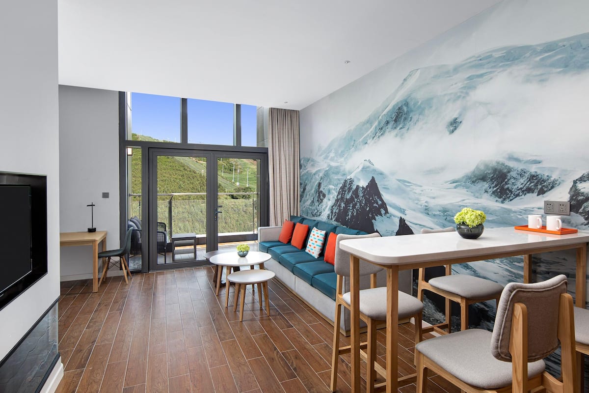 Una habitación moderna con un amplio ventanal, un sofá, una mesa de comedor y un mural con paisaje de montaña.