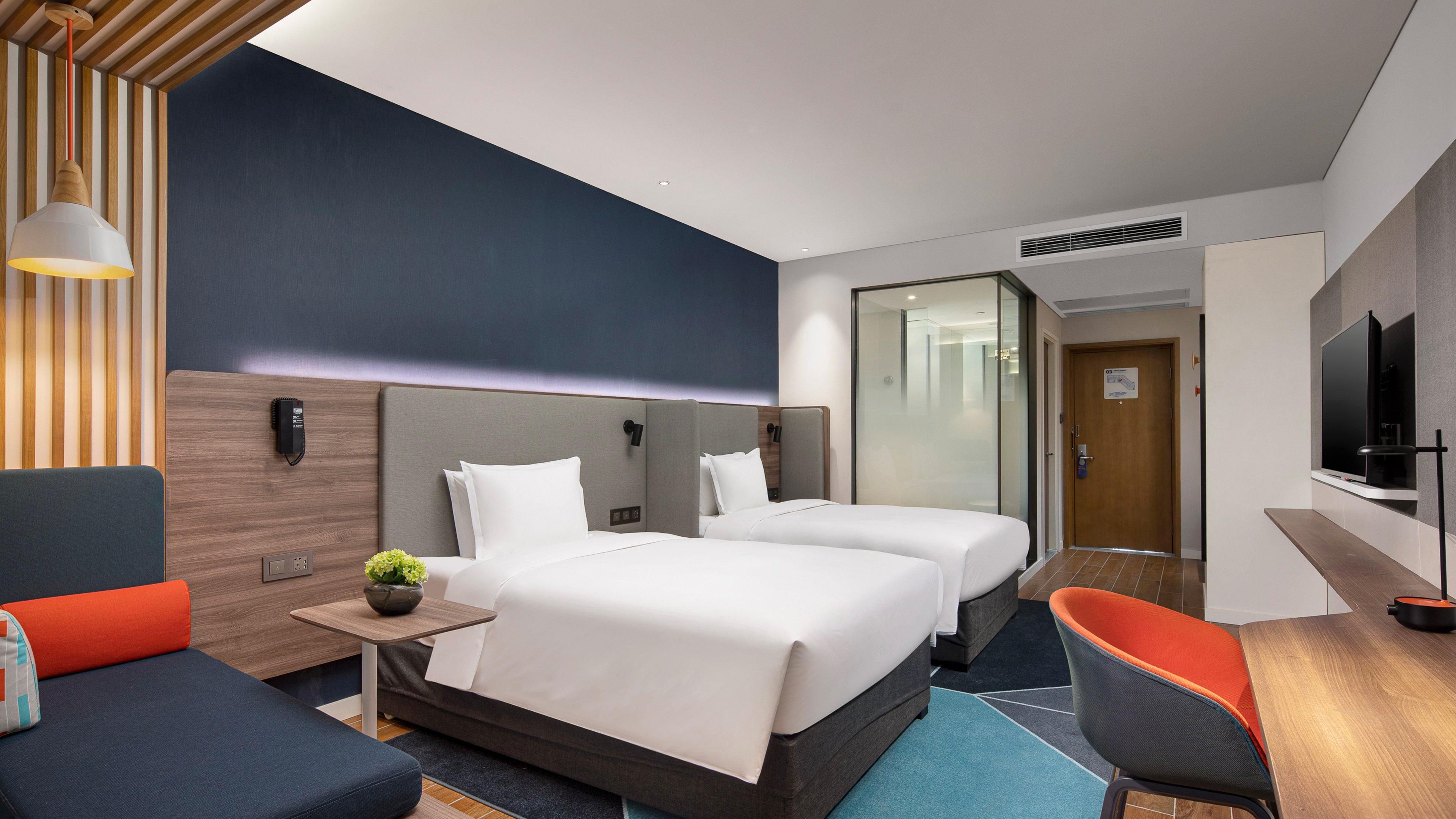 A hotel room with two beds, a desk, a TV, and a seating area with a plant.