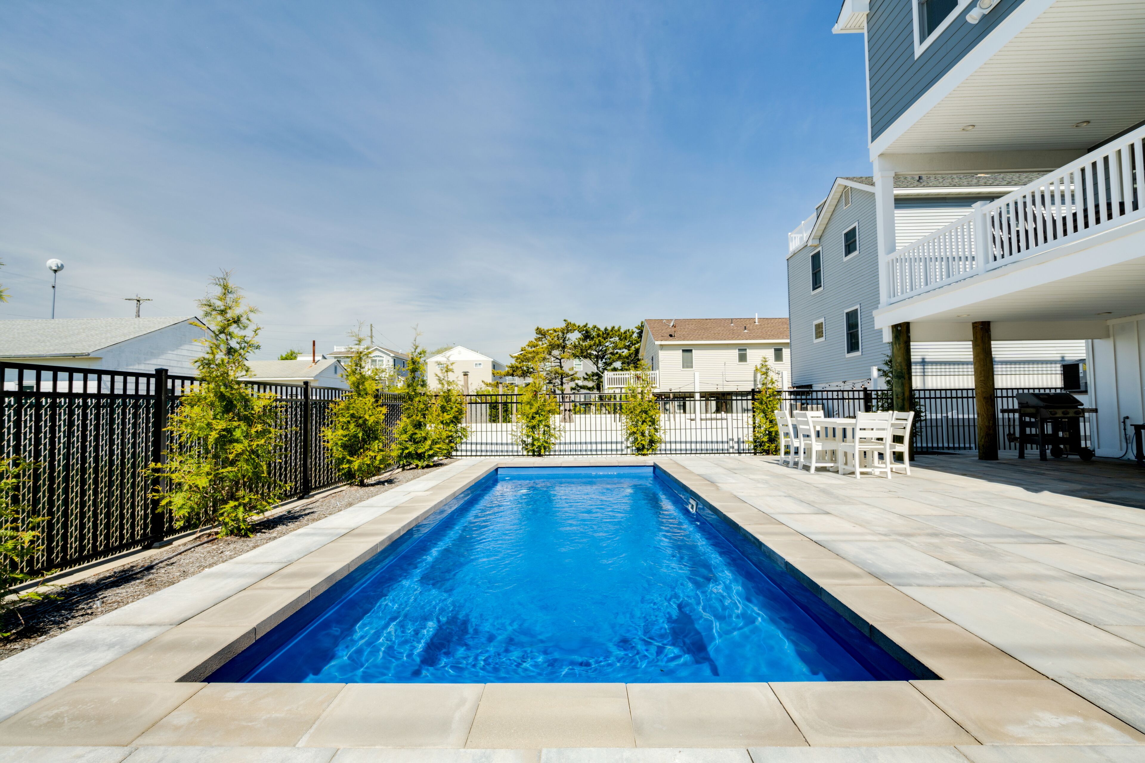 A rectangular swimming pool with a tiled deck, surrounded by a black fence and small trees.