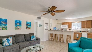A living room with a gray sofa, a teal armchair, a glass coffee table, and a ceiling fan. There are three paintings on the wall, a kitchen with wooden cabinets, a microwave, and a dishwasher.