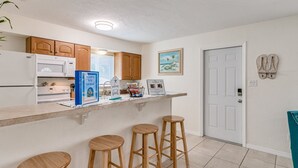 A kitchen with wooden cabinets, a white refrigerator, a microwave, and a coffee maker. There are three wooden stools and a framed welcome sign on the counter.