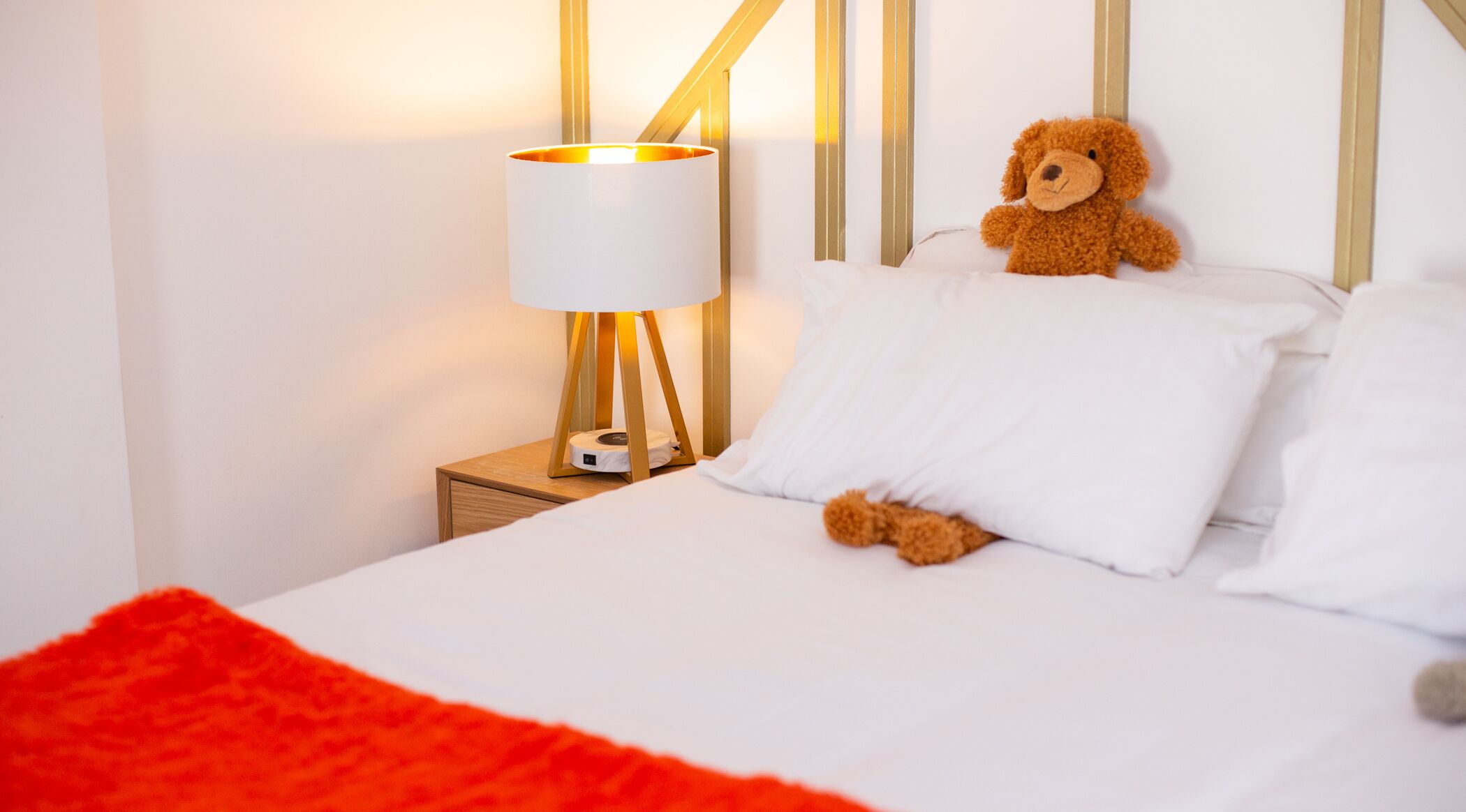 A neatly made bed with white linens and a red throw blanket, a bedside lamp with a white shade, and a teddy bear on the pillows.