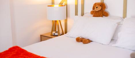 A neatly made bed with white linens and a red throw blanket, a bedside lamp with a white shade, and a teddy bear on the pillows.