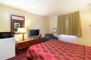 Habitación de hotel con cama, escritorio con televisor, microondas, lámpara, silla y ventana con cortinas.