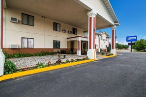 Un hotel con entrada cubierta, unidades de aire acondicionado, un banco y un área ajardinada con plantas y grava.