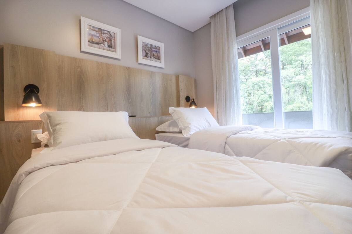 A hotel room with two beds, a wooden headboard, wall-mounted lamps, and a window with curtains.